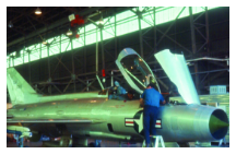 Technicians work on a silver MiG-21 fighter jet inside a large aircraft hangar. One technician stands on a ladder near the open cockpit. The aircraft displays a U.S. insignia.